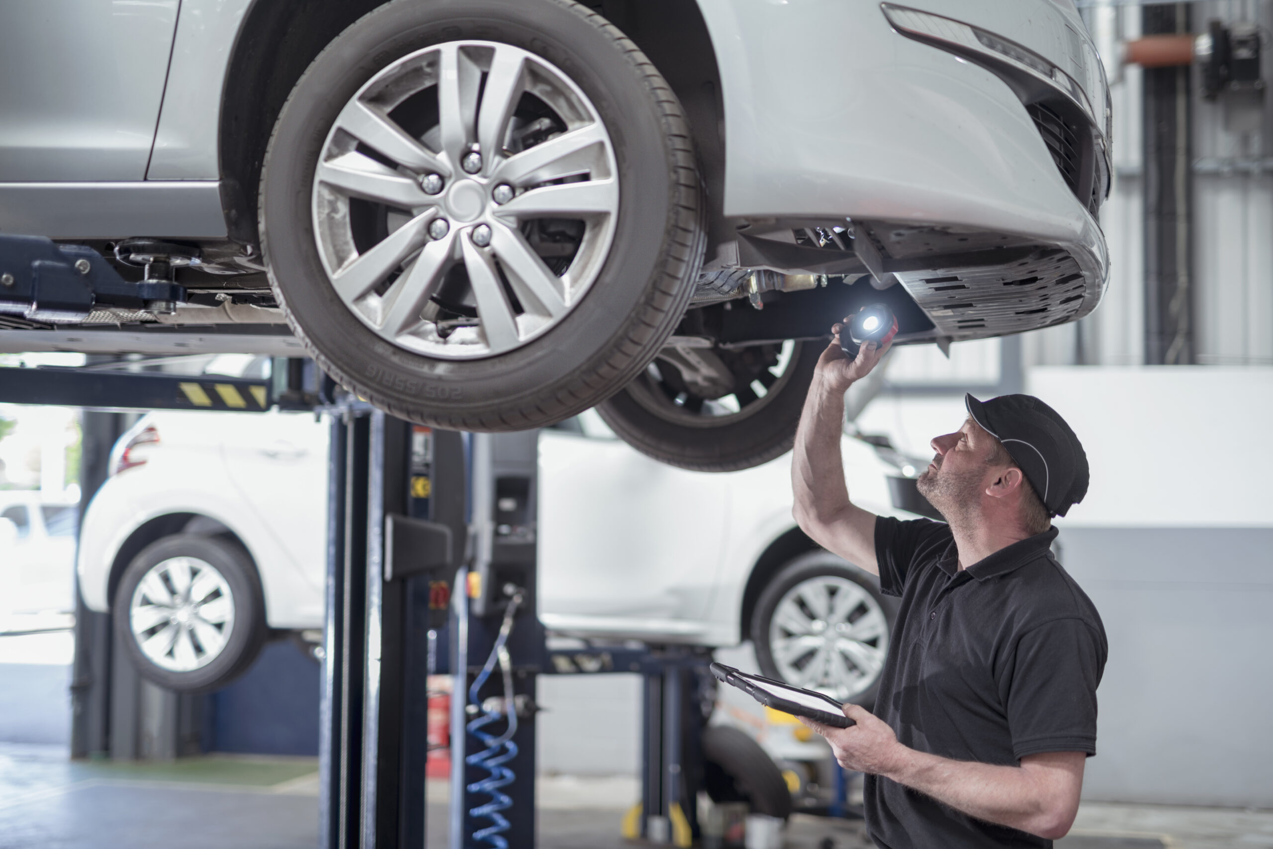 Mechanic inspecting the underside of a car with a flashlight and tablet.
